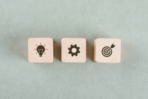 3 wooden blocks. Block 1 has a light bulb printed on it. Block 2 has a gear. Block 3 has a target. All 3 combined to denote the tools of project management.